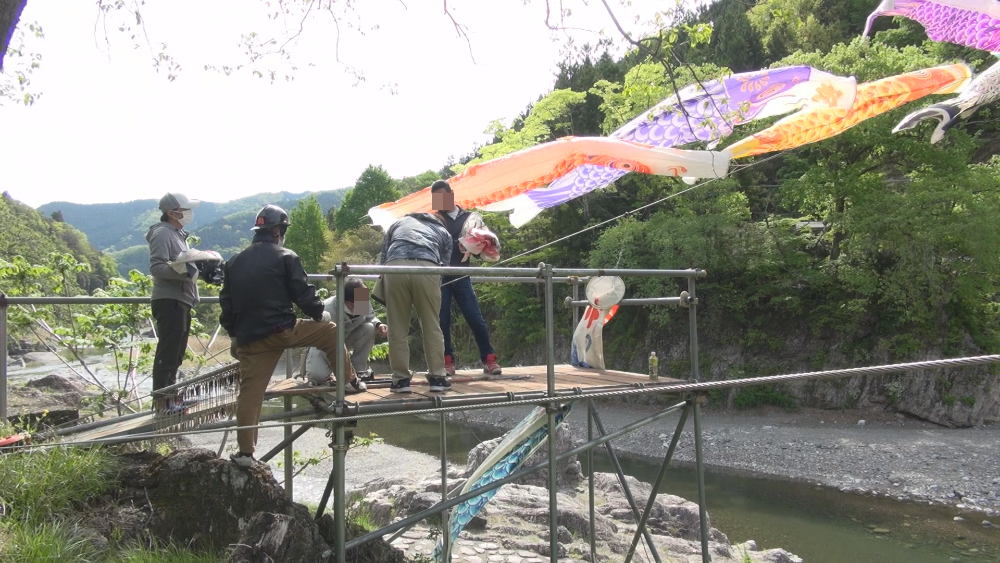 川辺でワイヤーに大きな鯉のぼりをかける人々