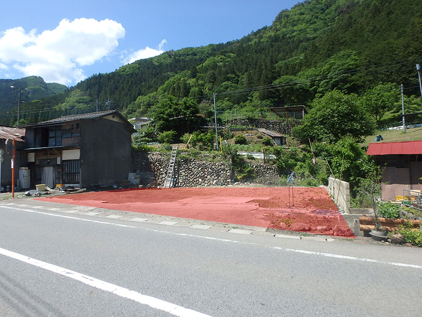 後方には山があり、左側には2階建ての建物と石が積み上げられた石垣が見え、道路沿いの一角が更地になっている土地の写真