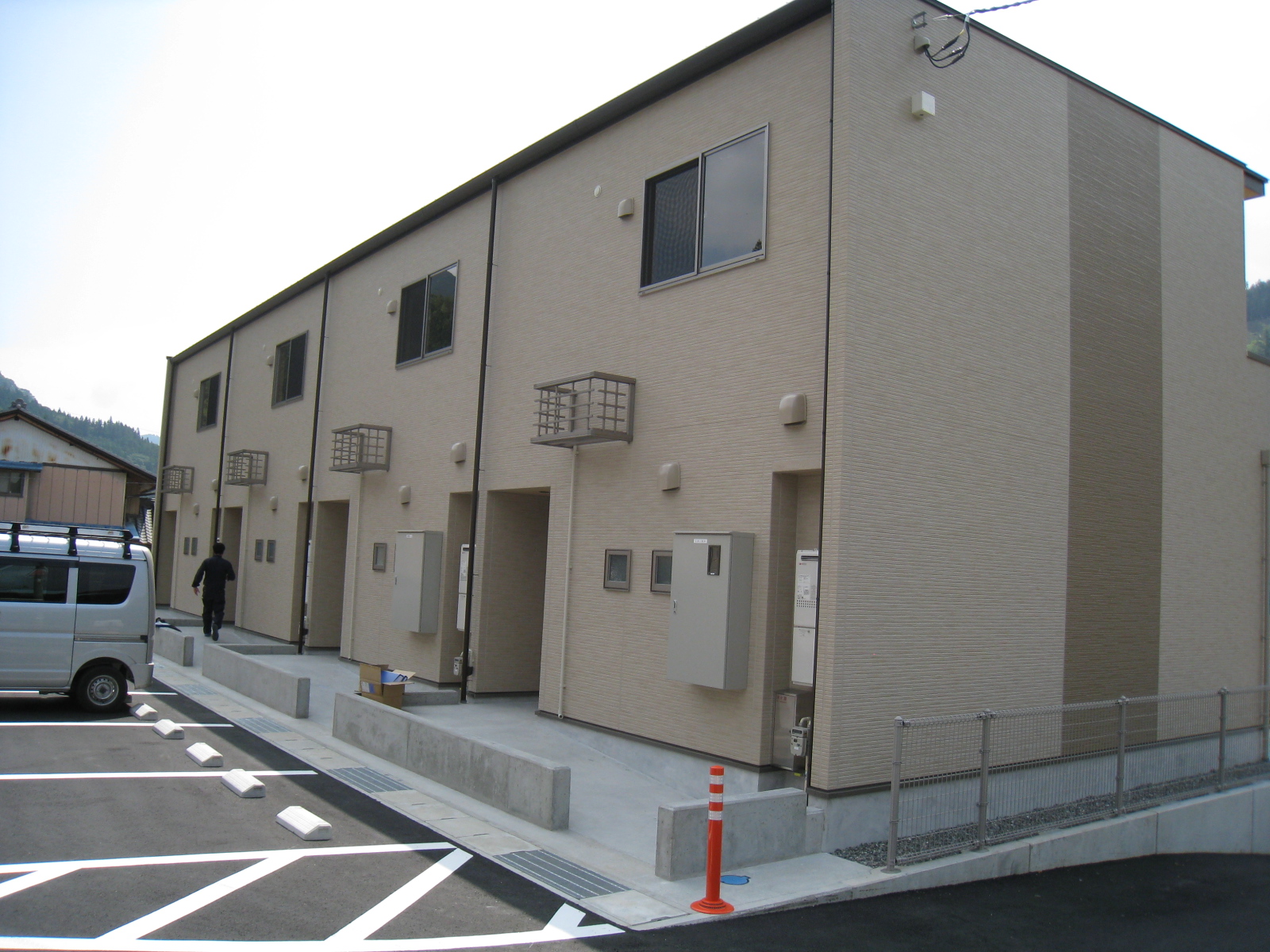 駐車スペースが建物前にあり、薄茶色を基調とした壁と、2階に長方形の窓が1つある木造2階建てのまちなみ団地A棟の写真