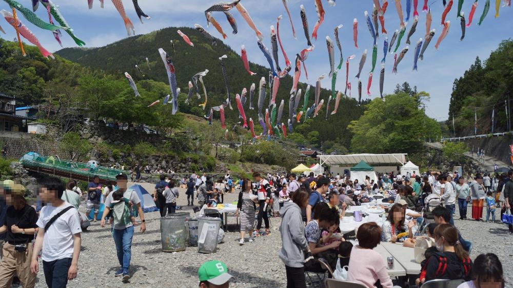 山間にかけられたたくさんの鯉のぼりの下の川辺で多くの人々が思い思いに過ごしている様子。